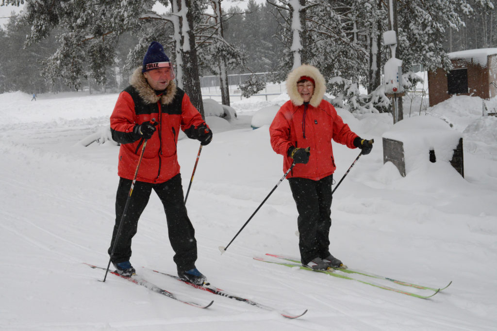 Mies ja nainen hiihtävät perintesellä tyylillä. Taustalla lumisia mäntyjä. 