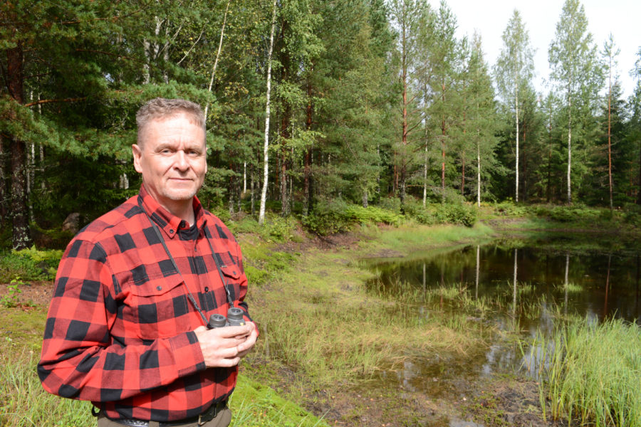 Metsästäjä lammen rannalla kiikarit kädessään.