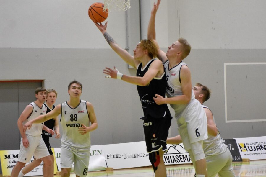 Mustapaitainen pelaaja nostaa koripalloa koriin, samalla kun valkopaitaiset pelaajat yrittävät estää.