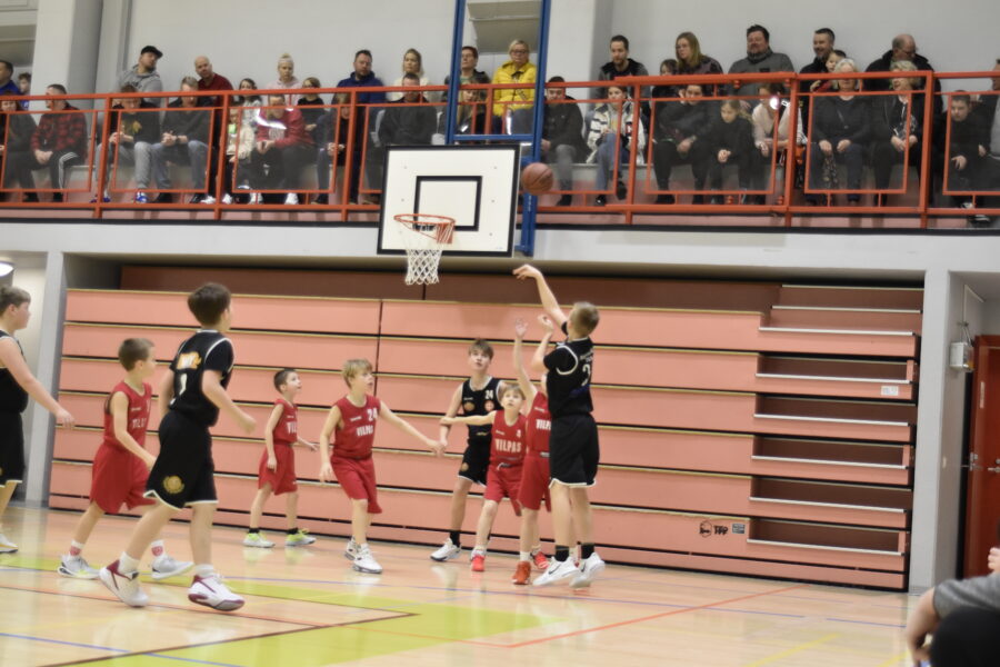 Nuori pelaaja mustassa paidassa yrittää koria yleisön edessä, punapaitaset puolustavat.